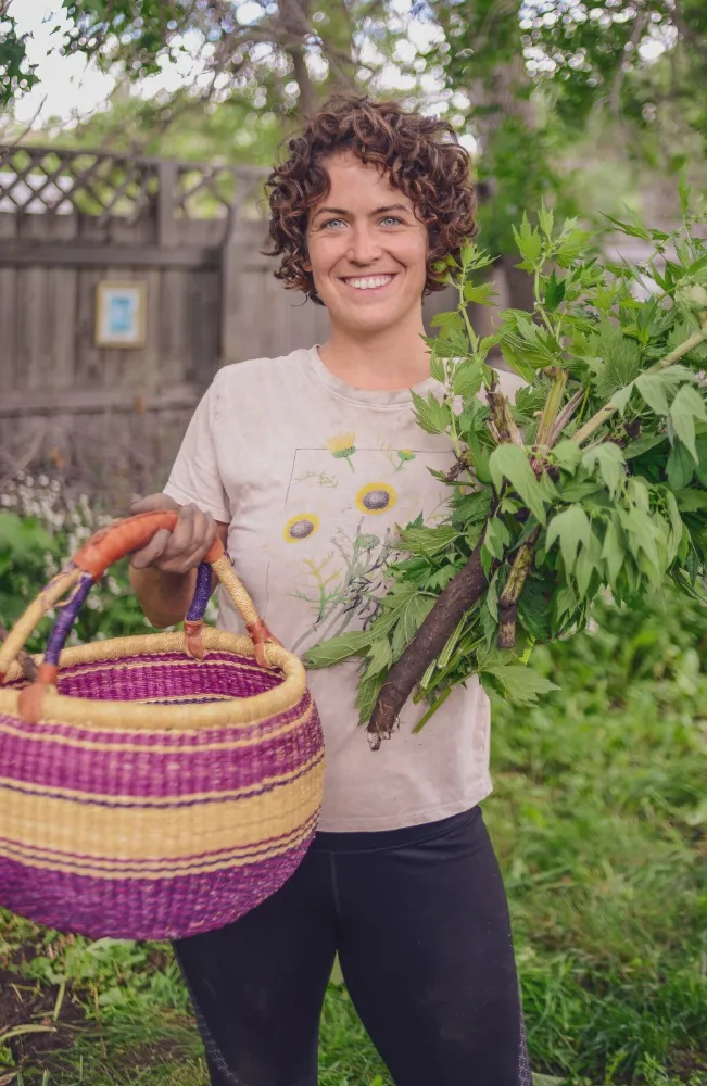 Student holding harvested herbs and a woven basket in a garden